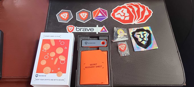 A "Crypto Stick - 6-Pack" hardware wallet product is displayed alongside its orange "SECRET RECOVERY SHEET," a Brave sticker, and various stickers including a lion logo and a holographic crypto design.