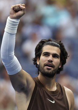 A male tennis player with dark hair and a beard, wearing a white sleeve on his raised arm and a brown Nike tank top, celebrating with a clenched fist in front of a blurred crowd.