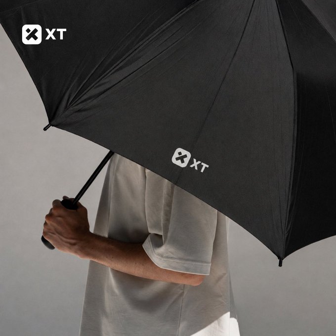 A person in a light-colored shirt holds up a black umbrella branded with the "XT" logo, visually reinforcing the post's message that XT provides protection in any weather as part of the #XTexchange campaign.