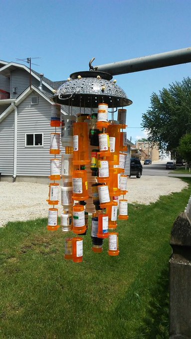 A hanging wind chime made from empty orange prescription pill bottles dangles outside a house, its cluttered strands of medication containers evoking the chaos of overmedication. The sculpture visually embodies the post's warning that "to defeat a monster u must become a monster," suggesting that the very pharmaceuticals meant to treat illness can themselves become a monstrous force when accumulated beyond control.