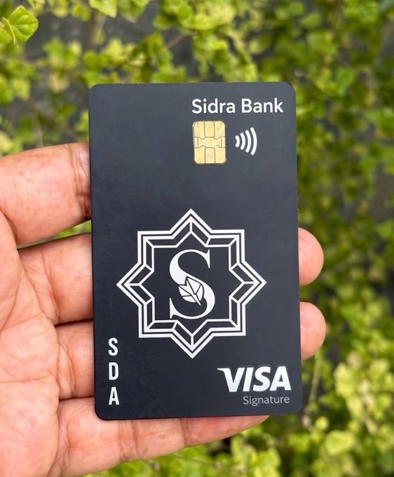 A hand holds a matte black Sidra Bank Visa Signature debit card featuring a gold EMV chip, contactless symbol, official Sidra emblem with an "S" and leaf design, and Visa Signature branding, set against a backdrop of green foliage—showcasing the premium new card described in the accompanying post.