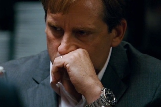 A man in a dark suit sits with his hand covering his mouth, wearing a silver wristwatch, deep in thought or frustration. His pensive expression mirrors the disbelief of the joke about Robinhood prioritizing its own blockchain over listing "fartcoin."