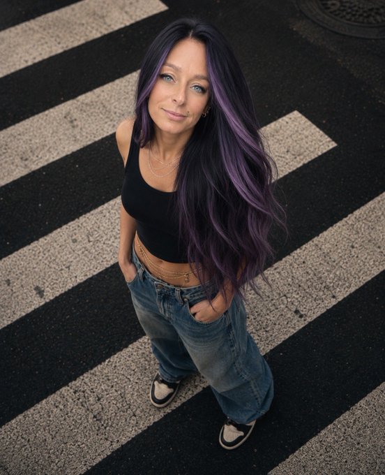 An overhead photograph of a young woman with long dark hair featuring purple highlights, wearing a black crop top, blue jeans, and sneakers, standing on a black and white striped crosswalk.