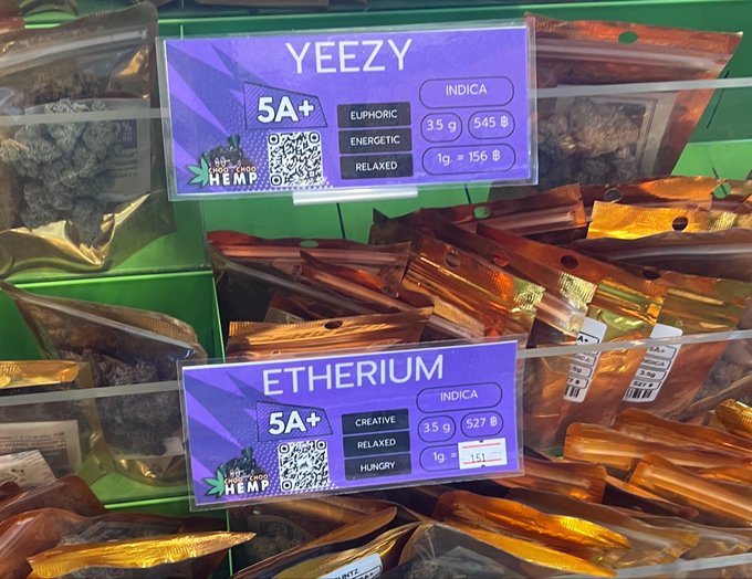The image shows a display of various bags of hemp products, including bags of hemp buds and hemp chocolate. There are two purple tags with white text that read "Yeezy", "Etherium", and "Indica". The tags also contain information about the product's weight, price, and quality. The background is green, which contrasts nicely with the purple tags and makes them stand out.
