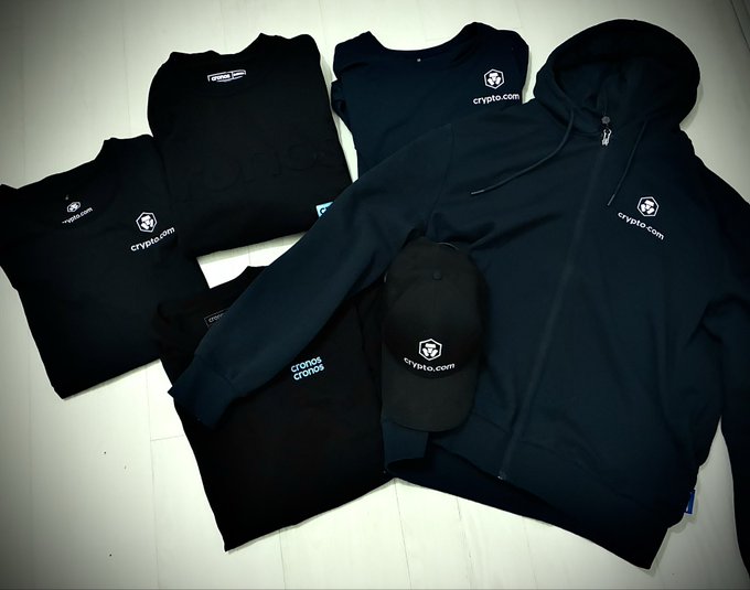 Flat lay of black Crypto.com branded apparel including t-shirts, hoodie, shorts, and cap arranged on a white tiled floor.
