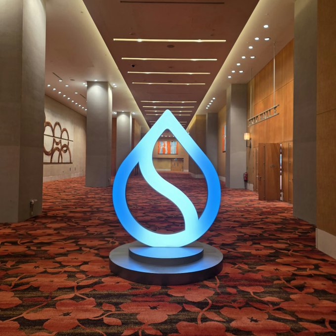 A large glowing blue Sui cryptocurrency logo sculpture positioned in the center of a hotel or convention hallway with patterned red carpet.