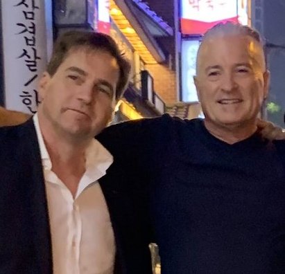 The image features two men standing next to each other, both wearing suits and smiling for a photo. They are positioned in front of a building that has neon lights illuminating its facade. The background includes a sign with Asian characters written in Japanese, which adds an interesting cultural context to the scene.