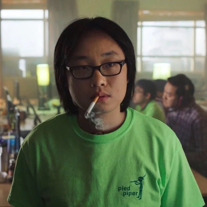 A man with glasses and long dark hair smokes a cigarette while wearing a bright green t-shirt featuring a 'pied piper' logo, set in a brightly lit office environment with other people working in the background.