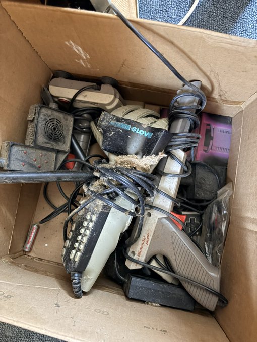 A cardboard box filled with dusty, tangled retro video game controllers and power supplies, including a 'Power Glove' and a Nintendo-branded accessory.