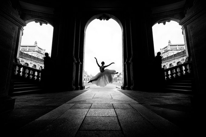In this black and white photo, there's a person dancing elegantly on a floor with an arched doorway in front of them. The door appears slightly open, and the scene suggests that they are either leaving or entering through the doorway. The architecture of the building is noteworthy as it seems to be part of a historical site like Dresden, Germany. The caption reads "GM Cabriole I don' t just photograph ballerinas, I also Photograph architecture. This place is in Dresden, and we see a city that was completely destroyed during World War II and has been wonderfully rebuilt". It's clear that the photo captures not only the dancer but also the grandeur of the building they're standing in front of.