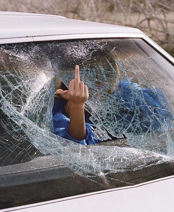 A person's hand giving a middle finger gesture through a shattered car windshield, with the spiderweb cracks clearly visible across the broken glass.