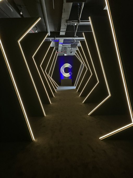 A dark indoor corridor featuring geometric black walls outlined with bright white LED light strips, leading toward a glowing blue 'C' logo at the end.