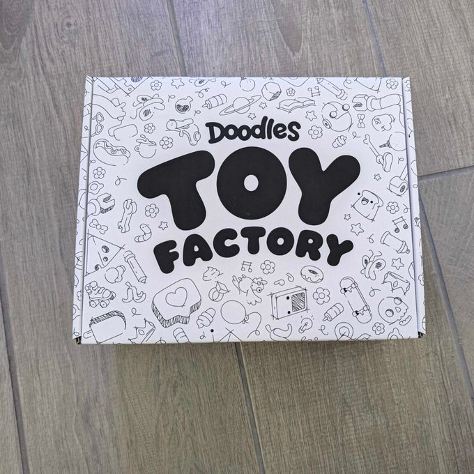A white package labeled "Doodles Toy Factory" sits on a wooden floor, surrounded by playful doodle illustrations of toys, animals, and whimsical characters. The colorful, cartoon-style box is perfectly suited for an unboxing video, hinting at the exciting collectibles awaiting inside.