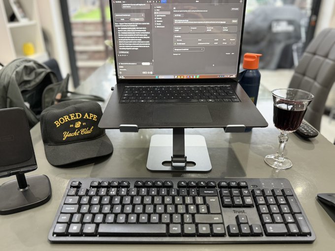 A desk setup featuring a laptop on a stand running an AI chat interface, a Bored Ape Yacht Club cap, a Trust keyboard, a phone stand, and a glass of dark liquid.