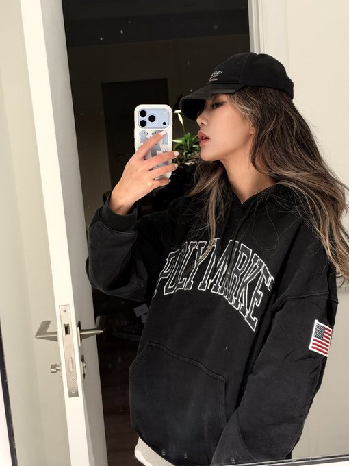 A woman in an oversized black "POLYMARKET" hoodie and black cap takes a mirror selfie with an iPhone, with an American flag patch on the sleeve. The image fits the prompt perfectly as it showcases branded Polymarket merchandise being modeled.