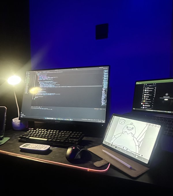 A nighttime coding setup featuring a monitor displaying code, a tablet with a sketch of cartoon characters, a laptop, mechanical keyboard, mouse, and a warm desk lamp against a blue-lit wall.