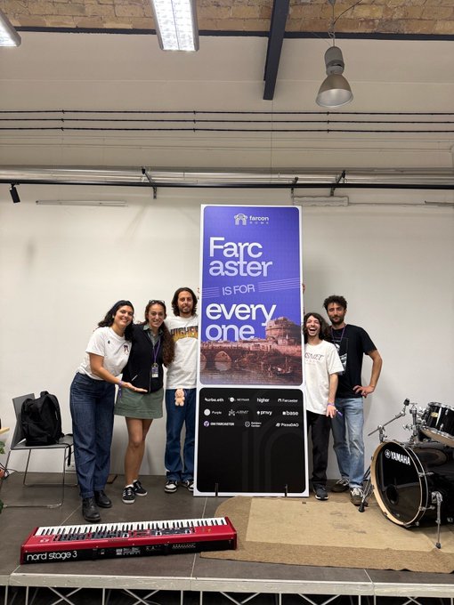 Five people posing together behind a banner that reads "Farcaster is for everyone" at a venue with musical equipment including a Nord Stage 3 keyboard and a Yamaha drum set.