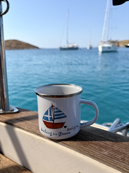 A white enamel mug reading "Sailing in Greece" with "Mocohima" branding sits on a boat's wooden railing, overlooking turquoise waters with sailboats anchored near a distant island—a perfect backdrop for the post's grateful message of good vibes and island daydreams.