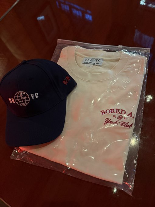 Bored Ape Yacht Club (BAYC) merchandise displayed on a wooden surface, featuring a navy cap and cream t-shirt with the "BAYC" logo and "Bored Ape Yacht Club" branding, still sealed in their plastic packaging—a new acquisition for the home in Hong Kong.