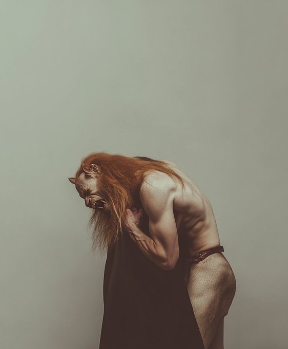 An artistic, muscular figure with long red hair and horns bends over against a pale background, wearing only dark straps.