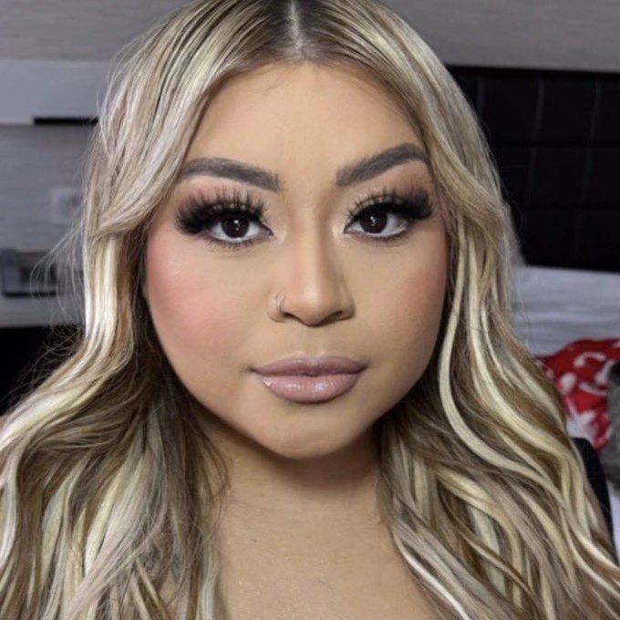 A close-up portrait of a woman with long wavy blonde hair, heavy eye makeup, false lashes, and a small nose ring.
