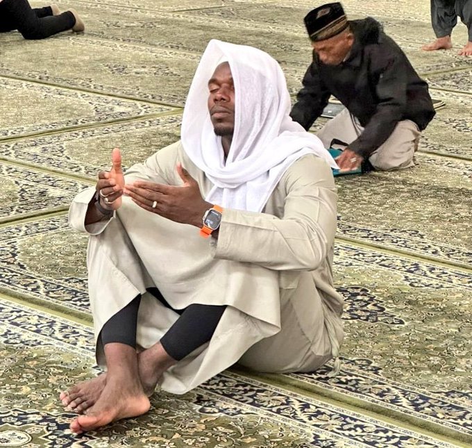 A Muslim man with a white head covering sitting on a prayer rug in a mosque, hands raised in prayer position.