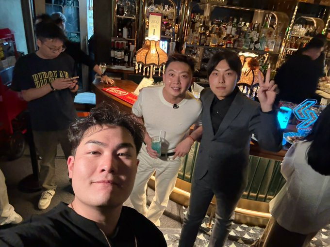 Three men pose for a selfie in a bar setting, with the man on the right making a peace sign, while another patron in a 'BURLBURN' shirt checks his phone in the background.