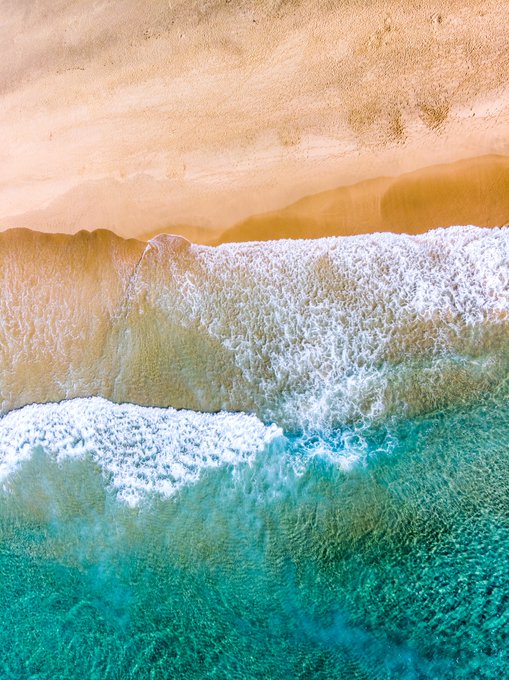 An aerial drone view of a pristine beach showing golden sand meeting turquoise ocean water with white foamy waves rolling onto the shore.