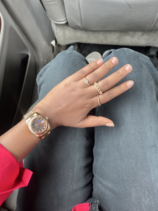 A close-up of a hand displaying a rose-gold watch and two rings (one square-cut, one eternity band) resting on dark denim-clad legs.