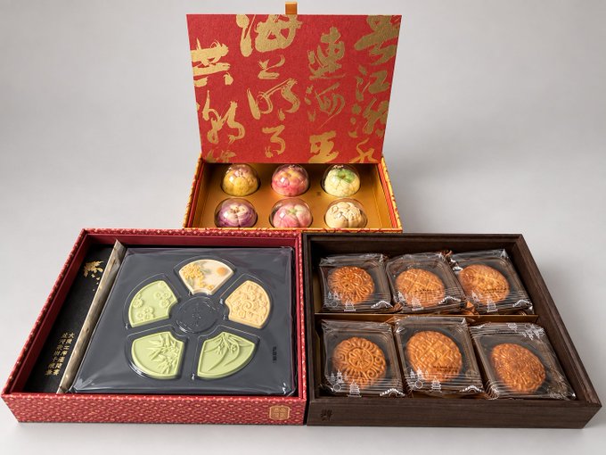 An open ornate red and gold box reveals an assortment of mooncakes and colorful pastries, with Chinese characters decorating the lid.