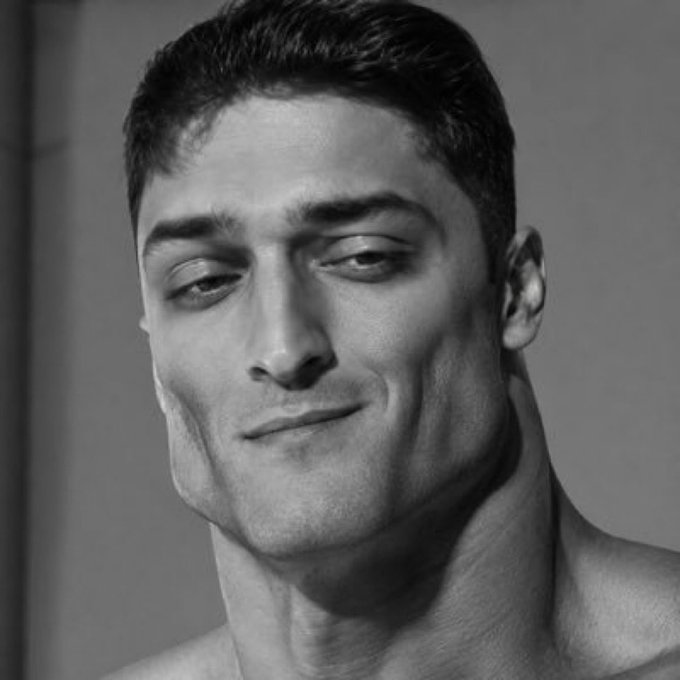 A black and white portrait of a man with short dark hair giving a subtle smirk, resembling a sculpture or painted artwork.