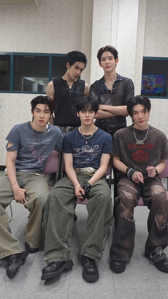 A group photo of five young men in stylish, edgy outfits including sleeveless tops, distressed denim, chains, and graphic tees, posing together in what appears to be a casual indoor setting.