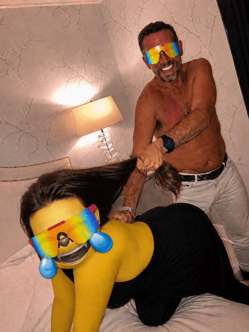 A photo of a shirtless man wearing rainbow sunglasses standing behind a woman on a bed, with the woman's face replaced by a laughing cat emoji with rainbow sunglasses and crying-laughing tears.