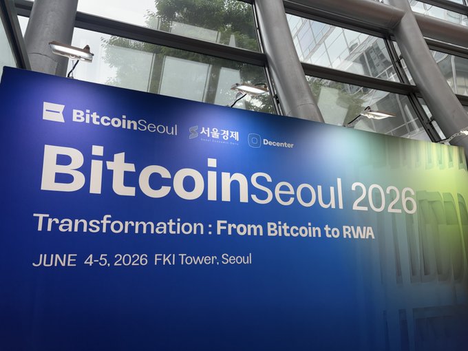The image shows a large blue sign with white text that reads "BITCOINSEoul 2026 Transformation : From Bitcoin to RWA". This suggests that there might be an event or conference related to the transformation of Bitcoin into a more widely accepted form of currency, possibly at the Seoul International Convention Center.

The background features a glass window and a building with columns, giving the impression that it is a convention center or large indoor space. The sign's position in front of this backdrop indicates its importance for the event being held within the venue.