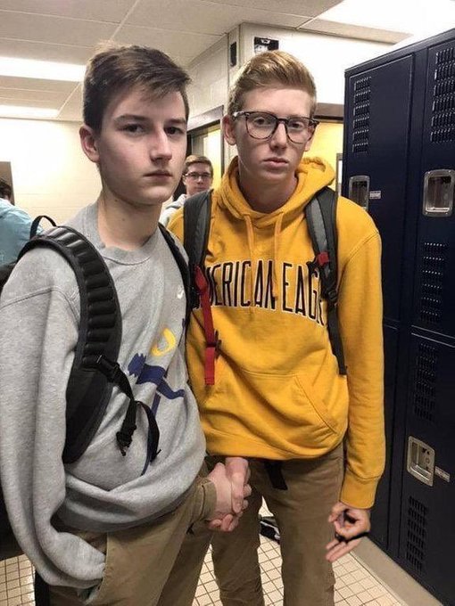 Two high school-aged boys stand together in a school hallway next to blue lockers, both wearing backpacks and serious expressions, with one in a gray hoodie featuring a crescent moon design and the other in a yellow "American Eagle" hoodie and glasses. The image humorously accompanies the meme about outperforming others in the meme coin market, jokingly framing these young students as savvy crypto traders whose youthful success leaves the poster humbled.