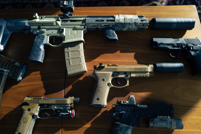 A tactical firearm collection is arranged on a wooden table, featuring an AR-style rifle equipped with a red dot optic and suppressor, alongside two semi-automatic pistols with attached lights and suppressors, a holstered handgun, and additional magazines—perfectly fitting the "Weapon Wednesday" theme. Visible markings include the "SureFire" branding on the weapon-mounted flashlight and the Beretta logo on the tan pistol.