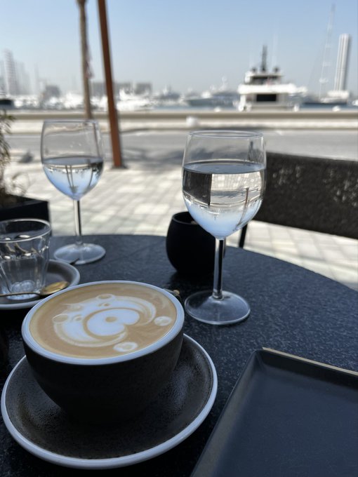 A cappuccino with bear-shaped latte art in a black cup on an outdoor cafe table, accompanied by glasses of water, with a marina featuring boats and yachts visible in the background.