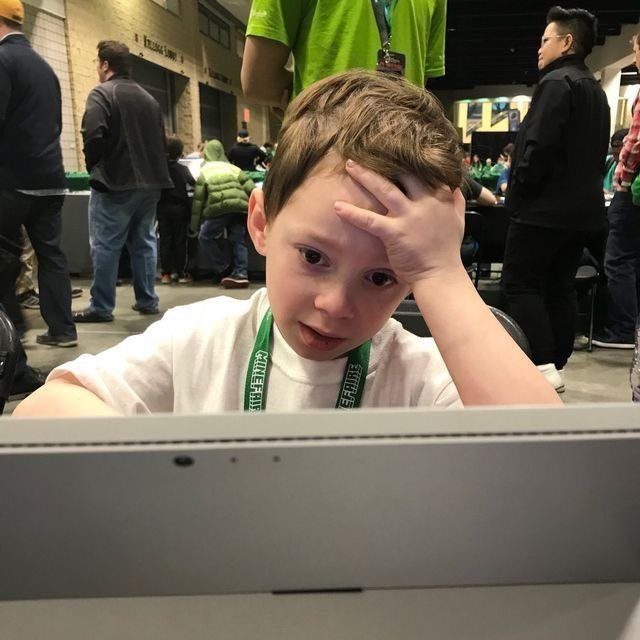 A young boy at what appears to be a convention event wearing a green lanyard, looking frustrated or overwhelmed while staring at a laptop screen with his hand on his forehead.