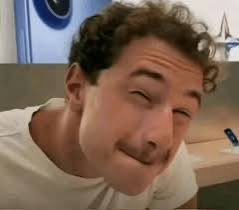 A man with curly brown hair and a mustache smiles widely with squinted eyes at the camera in an indoor setting.