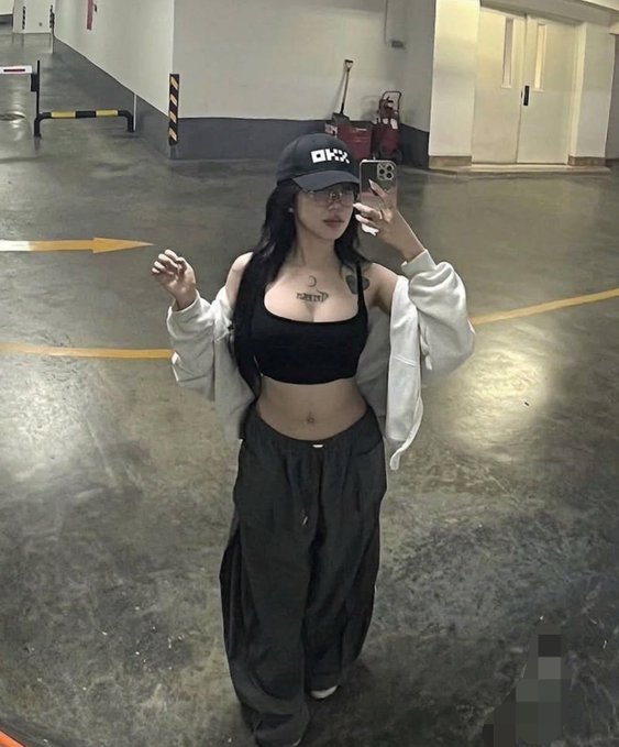 A young woman takes a mirror selfie in an underground parking garage, wearing a black crop top, oversized gray sweatpants, and a black cap with "DHX" text, paired with a cropped gray hoodie. Her relaxed, casual pose in the dimly lit garage matches the laid-back, weekend vibe of the "Happy Saturday Cuties" caption.