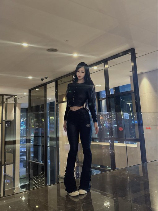A young woman with long black hair wearing a black top and black flared jeans posing in a modern lobby with reflective glass walls.