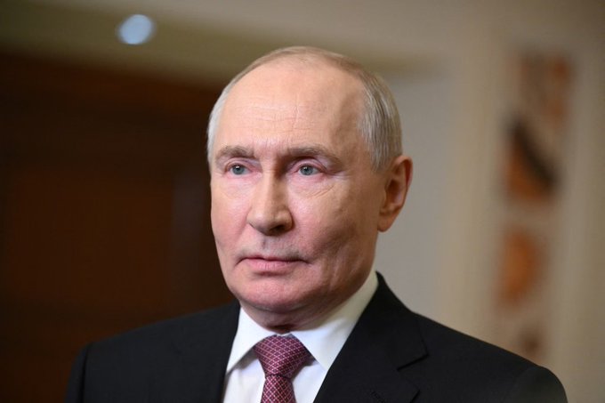 Portrait of Russian President Vladimir Putin wearing a black suit, white shirt, and maroon tie against a blurred interior background.