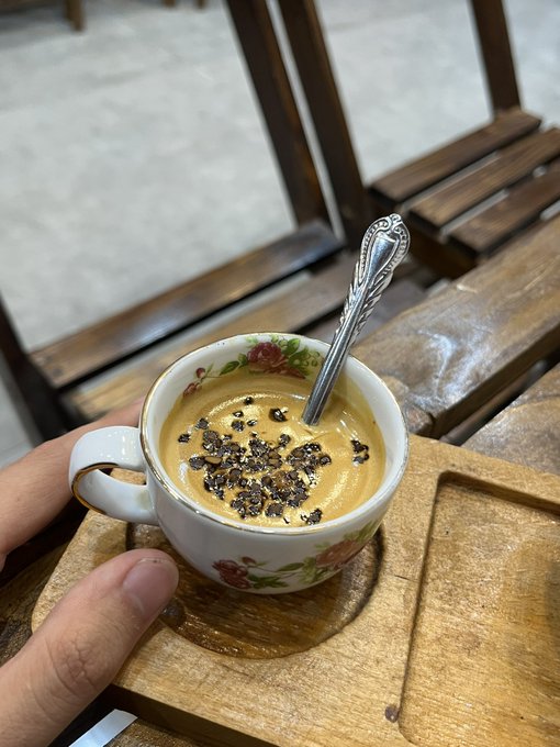 A hand holds a decorative floral teacup containing a coffee-like beverage topped with chocolate flakes, with a silver spoon resting inside, placed on a wooden tray and table.