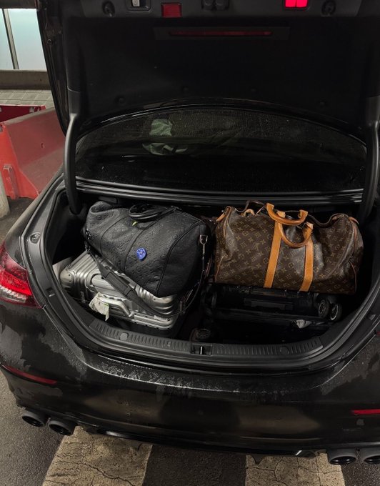 An open car trunk packed with travel luggage, including a brown Louis Vuitton duffel bag, a black backpack, a silver hard-shell suitcase, and what appears to be a folded stroller, suggesting preparation for a trip.