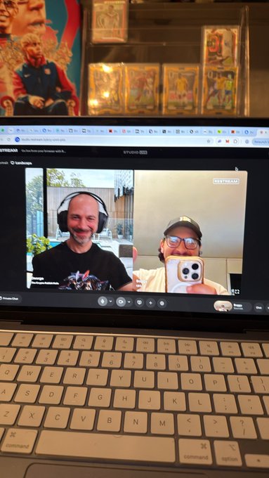 A laptop screen showing a video call between a bald man wearing headphones and another man in a cap taking a mirror selfie with an iPhone, with trading cards displayed on a shelf above the monitor.