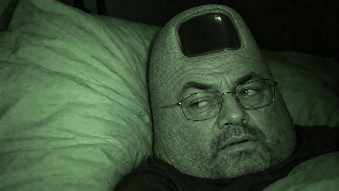 A green-tinted image of a man with glasses lying down on a pillow, with a surreal, smooth, helmet-like shape covering the top of his head that has a dark visor.