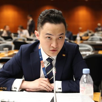 A man in a dark suit sits at a conference table, looking at the camera, with a nameplate and microphone in front of him and a water bottle to his side. The accompanying post about crypto billionaire Justin Sun suing his ex-girlfriend and her parents over a $4.5 million marriage gift is unrelated to this image, which appears to depict a professional conference setting and not necessarily Justin Sun.