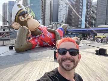 A selfie taken at ApeFest with the Bored Ape Yacht Club mascot statue, a large inflatable monkey character dressed in a colorful floral Hawaiian shirt and sunglasses, lounging on its side in an urban plaza. The image ties directly to the post's callout to BAYC fans by showcasing a memorable moment from an ApeFest event.