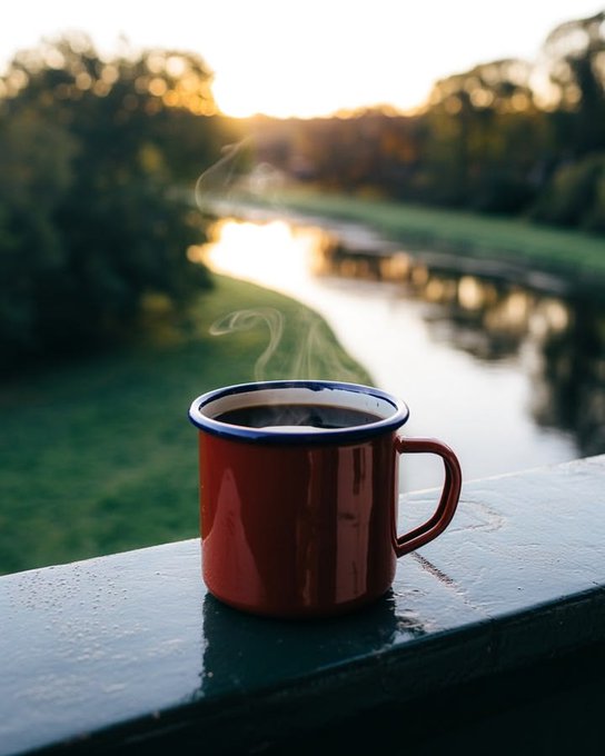 A steaming red enamel mug with a white rim sits on a green wooden ledge, overlooking a serene river and grassy bank at sunset.
