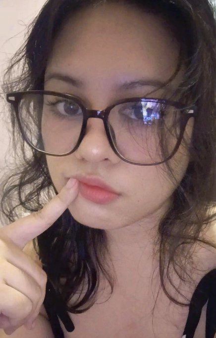 A close-up selfie of a young woman with dark wavy hair wearing black-rimmed glasses, touching her lips with her finger.